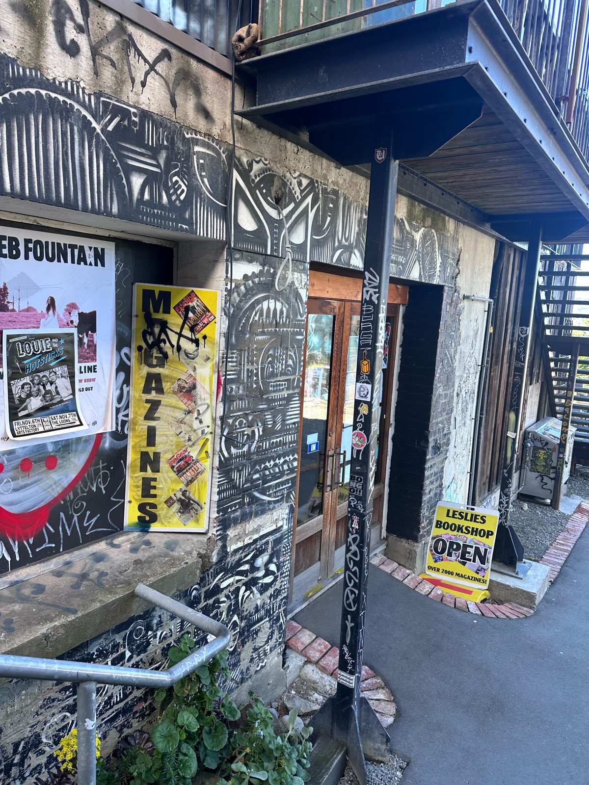 Leslie’s Bookshop entrance on London Street, Lyttelton, with a yellow MAGAZINES sign and black-and-white murals beneath an upstairs balcony