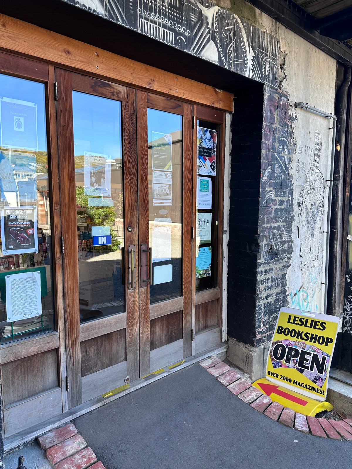 Wooden double doors of Leslie’s Bookshop in Lyttelton with a yellow Leslies Bookshop OPEN – over 2000 magazines sandwich board